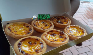 Egg tart Makau asli dengan kulit puff pastry renyah, custard telur kuning lumer, karamel hangus di atas, disajikan di piring putih