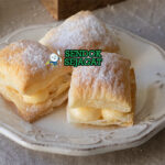 Miguelitos de La Roda siap saji: pastry puff renyah berlapis, diisi custard krim vanila lembut, ditaburi gula halus tebal, terlihat menggoda dan flaky