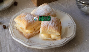 Miguelitos Siap Sajikan dengan Taburan Gula Halus Melimpah Miguelitos de La Roda siap saji: pastry puff renyah berlapis, diisi custard krim vanila lembut, ditaburi gula halus tebal, terlihat menggoda dan flaky