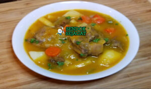 Mangkuk Soup Joumou Hangat Berisi Daging, Labu, Sayur, dan Pasta Kental Soup Joumou khas Haiti siap saji dalam mangkuk keramik, penuh potongan daging sapi empuk, labu kuning halus, wortel, kentang, kol, plantain, dan pasta dengan kuah kental gurih hangat