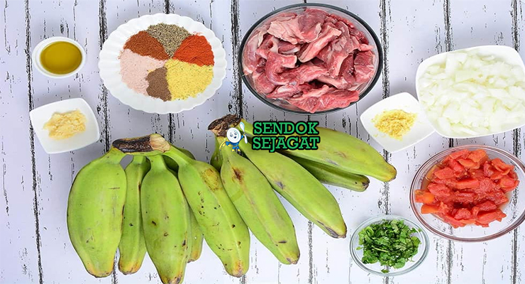 Bahan segar resep matoke ya nyama Uganda: pisang plantain matang, daging sapi potong dadu, tomat merah, bawang bombay, bawang putih, jahe, cabai merah, jinten, ketumbar, kunyit di atas meja dapur kayu sederhana