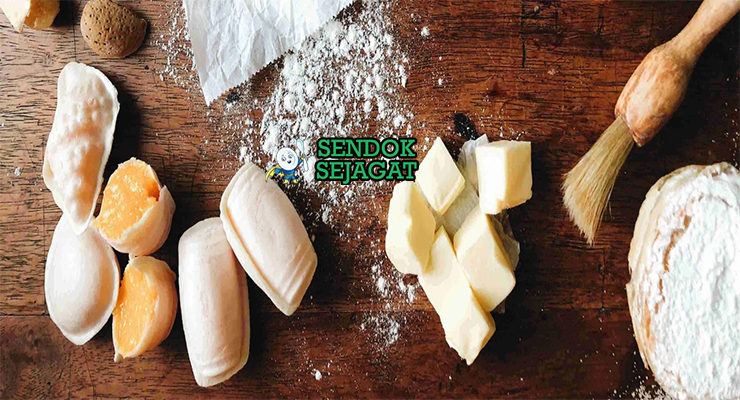 Bahan segar resep Pastéis de Tentúgal: kuning telur segar, gula pasir, tepung terigu protein tinggi, air, minyak zaitun, kayu manis batang, kulit lemon, mentega leleh di atas meja dapur kayu cerah