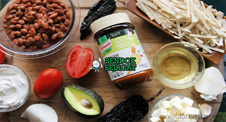 Bahan segar resep sopa tarasca Michoacán: kacang hitam kering, cabai guajillo kering, tomat matang, bawang bombay, bawang putih, oregano Meksiko, jinten, tortilla jagung, alpukat, keju cotija, krim asam di atas meja dapur kayu