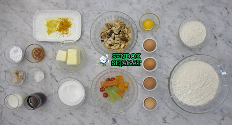 Bahan segar resep Bolo Rei: tepung terigu, gula pasir, telur segar, kismis, buah kandir campur, mentega tawar, kulit lemon, kulit jeruk, kayu manis bubuk di atas meja dapur kayu cerah