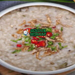 Bubur lambuk khas Malaysia siap saji: bubur beras kental lembut dalam mangkuk besar, tabur bawang goreng renyah, daun bawang iris, seledri, dan limau kasturi peras, terlihat gurih hangat dengan uap mengepul
