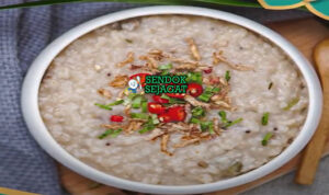 Bubur lambuk khas Malaysia siap saji: bubur beras kental lembut dalam mangkuk besar, tabur bawang goreng renyah, daun bawang iris, seledri, dan limau kasturi peras, terlihat gurih hangat dengan uap mengepul