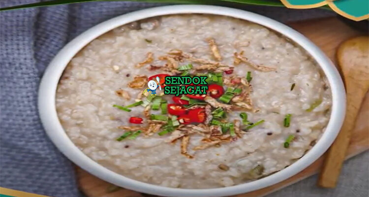 Bubur lambuk khas Malaysia siap saji: bubur beras kental lembut dalam mangkuk besar, tabur bawang goreng renyah, daun bawang iris, seledri, dan limau kasturi peras, terlihat gurih hangat dengan uap mengepul