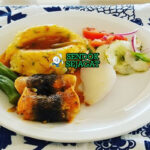 Cou-Cou and Flying Fish khas Barbados siap saji: bubur jagung okra kenyal lembut berbentuk bundar, ditumpuk flying fish goreng renyah keemasan, disiram saus tomat pedas manis, terlihat gurih segar dengan warna cerah tropis