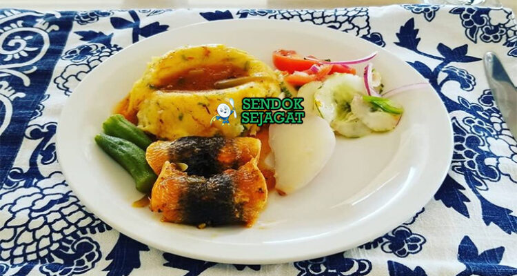 Cou-Cou and Flying Fish khas Barbados siap saji: bubur jagung okra kenyal lembut berbentuk bundar, ditumpuk flying fish goreng renyah keemasan, disiram saus tomat pedas manis, terlihat gurih segar dengan warna cerah tropis