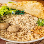 Cwie mie khas Malang siap saji: mangkuk mie kuning kenyal dengan topping ayam cincang gurih, bakso empuk, sawi segar, pangsit goreng kriuk, bawang goreng tabur, daun bawang iris, dan acar timun, terlihat hangat menggoda dengan minyak ayam wangi