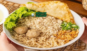 Cwie mie khas Malang siap saji: mangkuk mie kuning kenyal dengan topping ayam cincang gurih, bakso empuk, sawi segar, pangsit goreng kriuk, bawang goreng tabur, daun bawang iris, dan acar timun, terlihat hangat menggoda dengan minyak ayam wangi