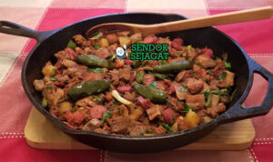 Discada khas Meksiko siap saji: wajan besar berisi tumisan daging sapi, chorizo, bacon, sosis, ayam, paprika merah-hijau, bawang bombay, tomat, cabai, terlihat gurih berapi dengan uap mengepul dan warna cerah