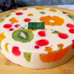 Jelly cake khas Australia siap saji: potongan irisan memperlihatkan lapisan jelly bening warna-warni (merah, hijau, kuning, biru), sponge cake lembut, dan buah segar terperangkap cantik, terlihat segar menggoda di piring putih