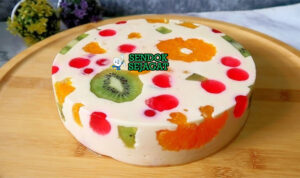Jelly cake khas Australia siap saji: potongan irisan memperlihatkan lapisan jelly bening warna-warni (merah, hijau, kuning, biru), sponge cake lembut, dan buah segar terperangkap cantik, terlihat segar menggoda di piring putih