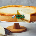 Käsekuchen Jerman Siap Disantap Potongan Käsekuchen khas Jerman bertekstur lembut creamy, permukaan kecokelatan halus, dasar biskuit renyah, disajikan di piring dengan garpu dan sedikit gula bubuk