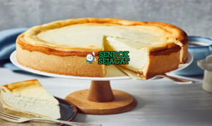 Potongan Käsekuchen khas Jerman bertekstur lembut creamy, permukaan kecokelatan halus, dasar biskuit renyah, disajikan di piring dengan garpu dan sedikit gula bubuk