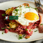 Labskaus khas Jerman utara siap saji: pure kentang lembut kental berwarna krem, daging kornet empuk, bit merah potong dadu, telur mata sapi kuning lumer di atasnya, herring rollmops acar di samping, terlihat gurih hangat menggoda
