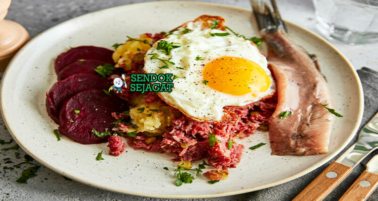 Labskaus khas Jerman utara siap saji: pure kentang lembut kental berwarna krem, daging kornet empuk, bit merah potong dadu, telur mata sapi kuning lumer di atasnya, herring rollmops acar di samping, terlihat gurih hangat menggoda