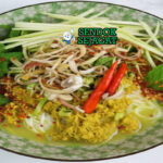 Nom Banh Chok khas Kamboja disajikan hangat dengan mie beras, kuah santan ikan kuning kehijauan, sayur mentah renyah, dan daun harum