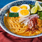 Ohn No Khao Swè khas Myanmar siap saji: mangkuk mie kuning kenyal dengan kuah santan kuning kental gurih, ayam suwir lembut, telur rebus, tauge segar, bawang goreng, daun bawang iris, dan kerupuk, terlihat hangat menggoda dengan uap mengepul