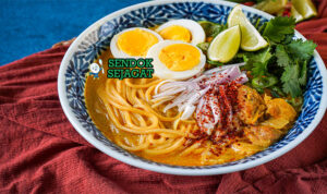 Ohn No Khao Swè khas Myanmar siap saji: mangkuk mie kuning kenyal dengan kuah santan kuning kental gurih, ayam suwir lembut, telur rebus, tauge segar, bawang goreng, daun bawang iris, dan kerupuk, terlihat hangat menggoda dengan uap mengepul