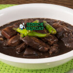 Pork dinuguan khas Filipina siap saji: sup darah babi hitam pekat gurih, potongan daging empuk, cabai hijau iris, dan kuah kental mengkilap, terlihat hangat menggoda dengan nasi putih