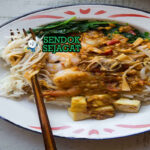 Satay bee hoon khas Singapura siap saji: mangkuk bihun lembut dengan kuah sate kacang manis gurih berwarna cokelat kental, udang panggang besar, tauge segar, telur rebus, tahu goreng, bawang goreng, dan limau kasturi, terlihat menggoda dengan uap hangat