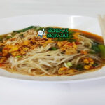 Shan noodles khas Myanmar siap saji: mangkuk mie beras kenyal dengan kuah tomat kacang tanah kental berwarna merah kecokelatan, daging cincang gurih, telur rebus, tauge segar, bawang goreng, kacang tanah cincang, daun bawang iris, dan jeruk nipis, terlihat hangat menggoda dengan uap mengepul
