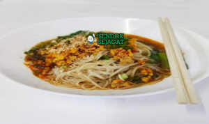 Shan noodles khas Myanmar siap saji: mangkuk mie beras kenyal dengan kuah tomat kacang tanah kental berwarna merah kecokelatan, daging cincang gurih, telur rebus, tauge segar, bawang goreng, kacang tanah cincang, daun bawang iris, dan jeruk nipis, terlihat hangat menggoda dengan uap mengepul