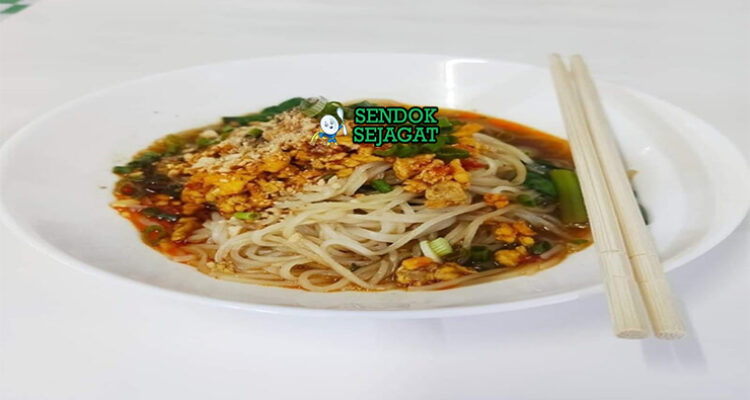 Shan noodles khas Myanmar siap saji: mangkuk mie beras kenyal dengan kuah tomat kacang tanah kental berwarna merah kecokelatan, daging cincang gurih, telur rebus, tauge segar, bawang goreng, kacang tanah cincang, daun bawang iris, dan jeruk nipis, terlihat hangat menggoda dengan uap mengepul