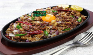 Sizzling pork sisig khas Filipina siap saji: daging babi cincang renyah di hot plate besi panas, bawang bombay segar, cabai rawit merah, jeruk nipis peras, uap mengepul dan bunyi sizzling menggoda