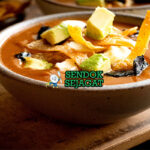 Sopa Tarasca khas Meksiko siap saji: mangkuk sup kacang hitam kental berwarna hitam kecokelatan, tabur tortilla goreng renyah, alpukat iris, keju cotija parut, krim asam, bawang merah, daun ketumbar segar, terlihat gurih pedas hangat dengan uap mengepul