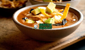 Sopa Tarasca khas Meksiko siap saji: mangkuk sup kacang hitam kental berwarna hitam kecokelatan, tabur tortilla goreng renyah, alpukat iris, keju cotija parut, krim asam, bawang merah, daun ketumbar segar, terlihat gurih pedas hangat dengan uap mengepul