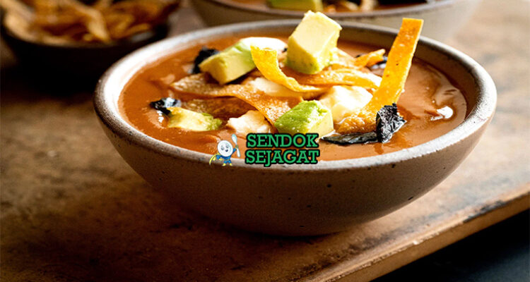 Sopa Tarasca khas Meksiko siap saji: mangkuk sup kacang hitam kental berwarna hitam kecokelatan, tabur tortilla goreng renyah, alpukat iris, keju cotija parut, krim asam, bawang merah, daun ketumbar segar, terlihat gurih pedas hangat dengan uap mengepul