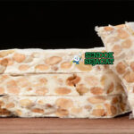Turrón khas Spanyol siap saji: blok nougat keras Alicante dengan almond utuh terlihat renyah, madu mengkilap, potong kotak di piring kayu, terasa manis dan crunchy