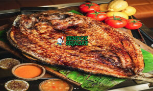 Masgouf khas Irak dengan ikan mas panggang terbuka renyah kecokelatan, asap kayu bakar, tampak gurih hangat menggugah selera