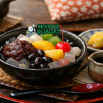 Anmitsu khas Jepang dengan kubus agar-agar kenyal, pasta kacang merah, ceri segar, kiwi, mandarin, dan sirup hitam disajikan di mangkuk kaca dengan es batu