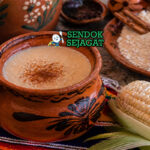 Minuman kental berwarna krem dalam cangkir dengan batang kayu manis dan uap hangat yang menggoda