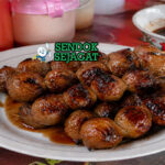 Bakso bakar khas Malang berwarna cokelat gelap mengkilap dengan lapisan saus kecap manis pedas, terlihat juicy dengan sedikit gosong di permukaan hasil panggangan arang, disajikan dalam tusuk sate dengan taburan daun bawang