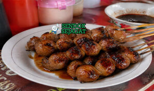 Bakso bakar khas Malang berwarna cokelat gelap mengkilap dengan lapisan saus kecap manis pedas, terlihat juicy dengan sedikit gosong di permukaan hasil panggangan arang, disajikan dalam tusuk sate dengan taburan daun bawang