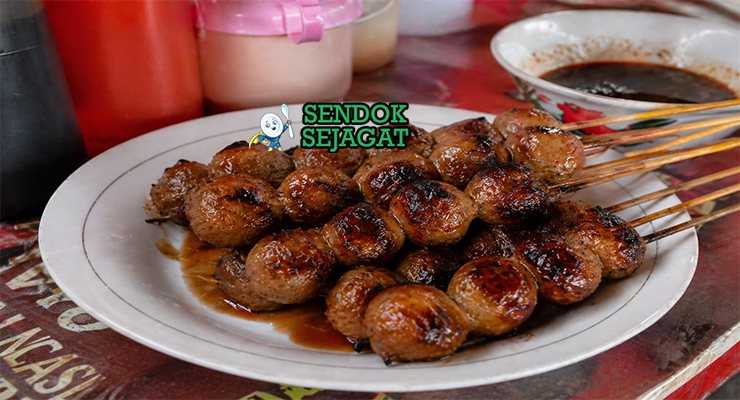 Bakso bakar khas Malang berwarna cokelat gelap mengkilap dengan lapisan saus kecap manis pedas, terlihat juicy dengan sedikit gosong di permukaan hasil panggangan arang, disajikan dalam tusuk sate dengan taburan daun bawang
