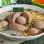 Bakso beranak berukuran besar dalam mangkuk dengan kuah kaldu bening, dibelah memperlihatkan bakso kecil di dalamnya, lengkap dengan mie, daun bawang, dan taburan seledri