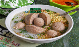 Bakso beranak berukuran besar dalam mangkuk dengan kuah kaldu bening, dibelah memperlihatkan bakso kecil di dalamnya, lengkap dengan mie, daun bawang, dan taburan seledri