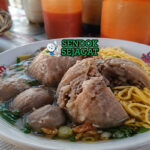 Bakso berwarna cokelat muda dengan potongan urat di dalamnya, disajikan dalam kuah bening dengan daun bawang dan bawang goreng