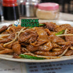 Hidangan beef chow fun dengan mie lebar berwarna cokelat mengilap, irisan daging sapi juicy, tauge segar, dan daun bawang dalam wajan panas