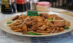 Hidangan beef chow fun dengan mie lebar berwarna cokelat mengilap, irisan daging sapi juicy, tauge segar, dan daun bawang dalam wajan panas