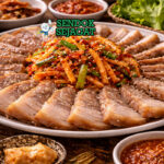Hidangan bossam Korea autentik dengan irisan daging empuk juicy, daun selada hijau kriuk, kimchi merah, dan mangkuk ssamjang pedas di piring putih yang menggugah selera