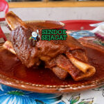 Potongan kaki babi chamorros de cerdo berwarna cokelat keemasan dengan tekstur empuk, disiram saus merah pekat, disajikan dengan jeruk nipis dan tortilla hangat