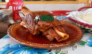 Potongan kaki babi chamorros de cerdo berwarna cokelat keemasan dengan tekstur empuk, disiram saus merah pekat, disajikan dengan jeruk nipis dan tortilla hangat