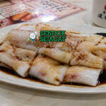 Gulungan chee cheong fun putih halus dengan tekstur licin disiram saus kecap khas, ditaburi wijen dan daun bawang, tampilan mengilap menggugah selera